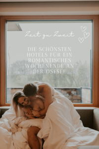 Romantisches Wochenende an der Ostsee für dich und deinen Liebsten | Das Bild zeigt ein Pärchen, dass sich verliebt im Bett kabbelt. Im Hintergrund ist ein großes Fenster mit Blick auf einen regnerischen Wald