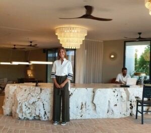 Eine junge Frau in der Uniform des Maslina Resorts steht vor der Rezeption und lächelt in die Kamera. Sie zeigt die nachhaltige Arbeitskleidung im Hotel Maslina Resort