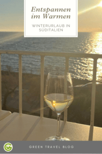 Auf dem Bild sieht man ein Glas mit Weißwein auf einer Veranda. Im Hintergrund sieht man das Meer bei Sonnenuntergang