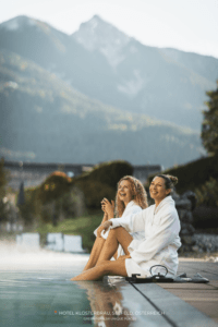 Zwei Frauen in weißen Gewändern sitzen am Pool des nachhaltigen Hotels Klosterbräu, lachen entspannt und genießen den Blick auf die Berge im Hintergrund. Die Stimmung ist heiter und fröhlich.