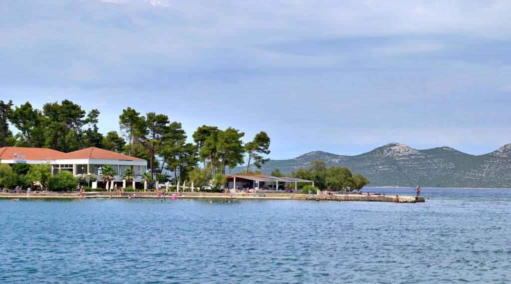 Coastal scene with a resort building amid pine trees, facing a calm, wide ocean. People gather by the shore. Hills are visible in the background, under a cloudy sky.