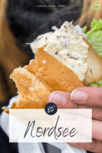 Nahaufnahme einer Hand, die ein Sandwich mit Frischkäse, Salat und Fisch hält. Der Textüberlagerung lautet „Kulinarische Reise zur Nordsee“ in eleganter Schrift.