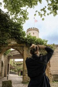 Person von hinten in schwarzer Kleidung fotografiert, die eine historische Burg mit Rundturm und Fahne betrachtet, umgeben von grünen Bäumen und einer Steinmauer mit Bogen