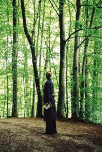 Frau in dunklem Mantel steht auf Waldweg im Teutoburger Wald und hält ein Buch in der Hand