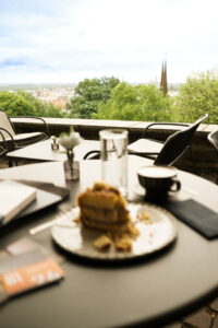 Tisch mit Kaffee, Glas Wasser und Stück Kuchen auf Terrasse mit Blick auf Stadt und Kirchturm im Hintergrund