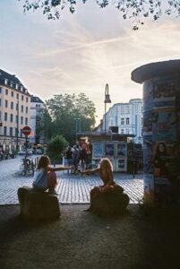 Zwei Personen sitzen auf großen Steinen und reichen sich an einem Kiosk in einer belebten Straße in Köln Getränke