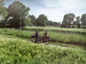 Zwei Personen fahren auf Fahrrädern auf einem schmalen Weg durch eine grüne, ländliche Landschaft mit Bäumen und Wiesen im Hintergrund