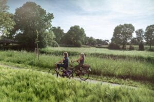 Zwei Personen fahren auf Fahrrädern auf einem schmalen Weg durch eine grüne, ländliche Landschaft mit Bäumen und Wiesen im Hintergrund