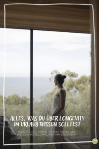 Frau im Bademantel steht vor großem Fenster mit Blick auf grüne Landschaft und Berge im Wellnesshotel mit Longevity-Angebot