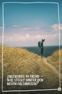 Wanderer mit Rucksack und Hut steht auf einem schmalen Pfad zwischen hohen Gräsern mit Blick aufs Meer unter blauem Himmel