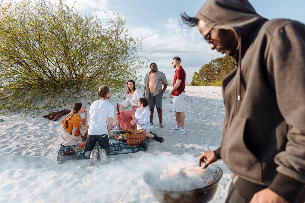 Gruppe von Leuten am Strand in den Dünen machen Picknick. Ein Mann steht im Vordergrund am Grill
