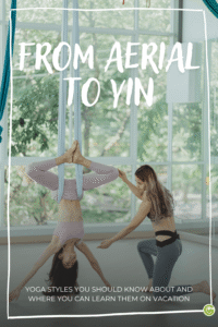 A woman hangs upside down in an aerial yoga hammock while another woman guides her, in a bright room with large windows and a view of greenery outside; text reads “FROM AERIAL TO YIN” and “YOGA STYLES YOU SHOULD KNOW—AND WHERE YOU CAN LEARN THEM ON VACATION”