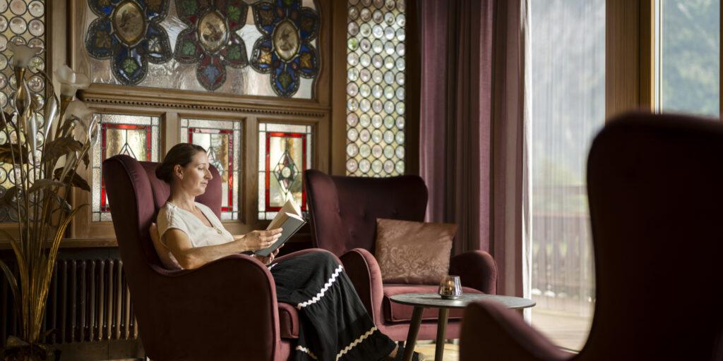 Hotel interior featuring natural materials. A woman sits in the lobby in front of the panoramic window overlooking the Allgäu.