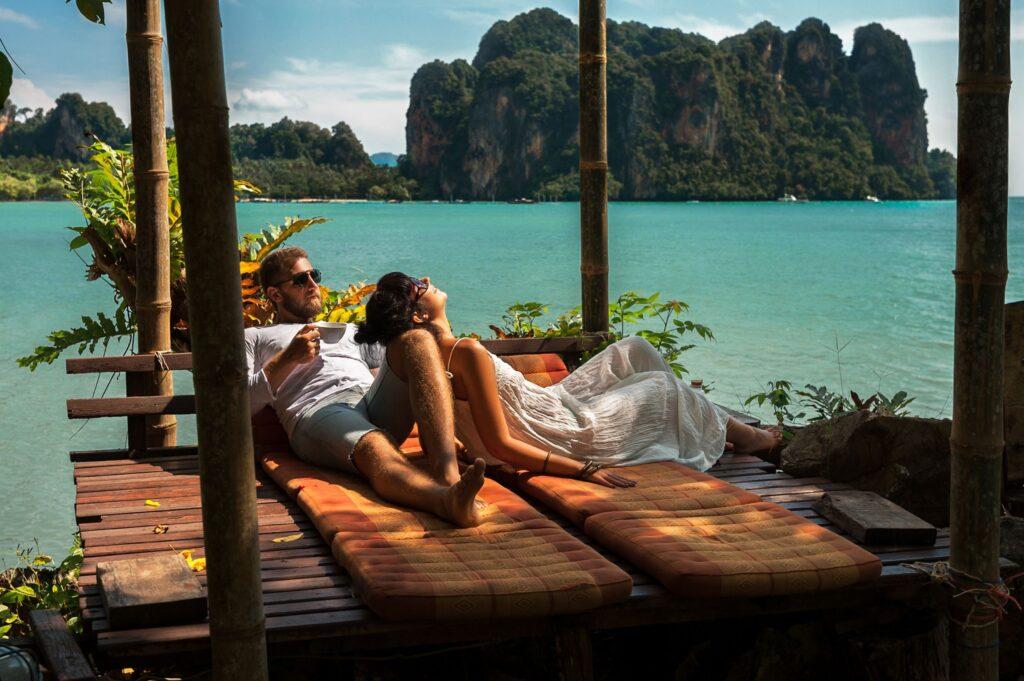 Paar in Thailand relaxt auf einem Pier mit Blick auf das türkiese Wasser und Felsen. Unbeschwert und bewusst Reisen
