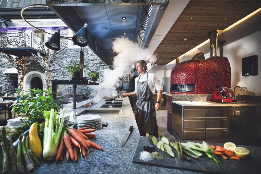 Koch in moderner Hotelküche mit dampfendem Herd, offenem Steinofen und frischem Gemüse auf Arbeitsflächen