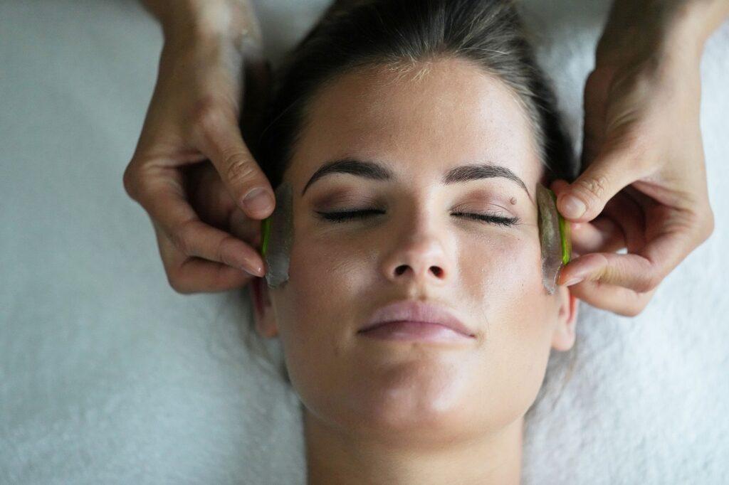 Frau mit geschlossenen Augen erhält Gesichtsmassage mit zwei Aloe-vera-Blättern an den Wangen